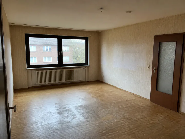 Leerer Wohnraum mit strukturierter Tapete, abgenutztem Holzboden, Heizkörper unter dem Fenster und brauner Tür mit Glaseinsatz
