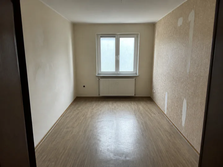 Kleines leeres Zimmer mit Heizkörper unter dem Fenster, hellem Boden in Holzoptik und ausgebesserten Stellen an der Tapete
